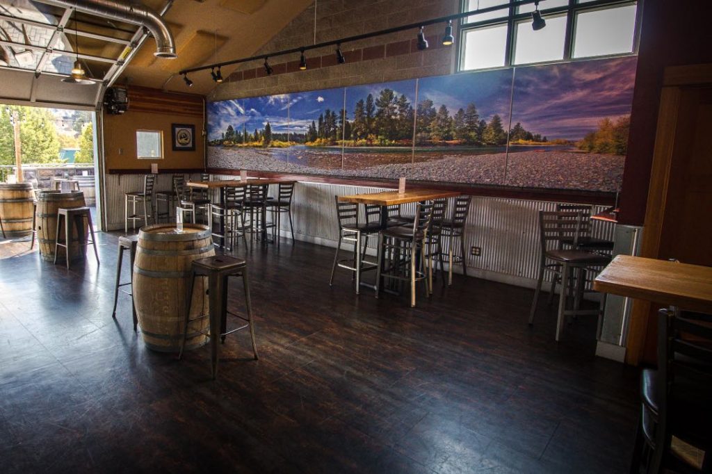 Brewery interior with modern high top tables, a barrel with arranged around it, and a mural of a mountain landscape on the wall.