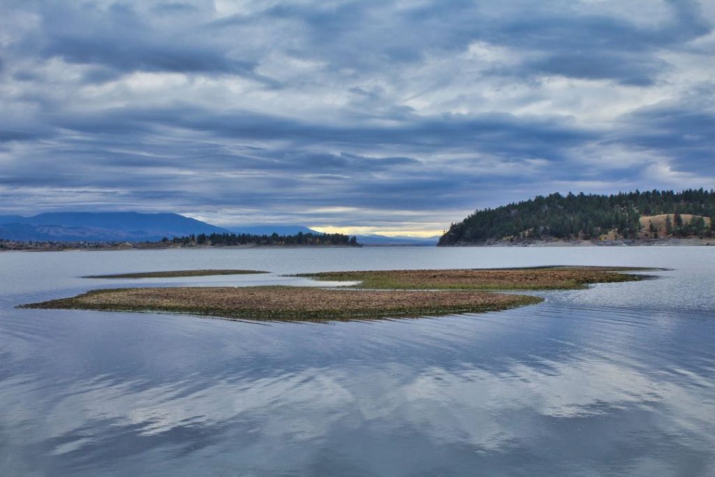 Dark clouds hang over a glassy lake with a few forested hills in the background.