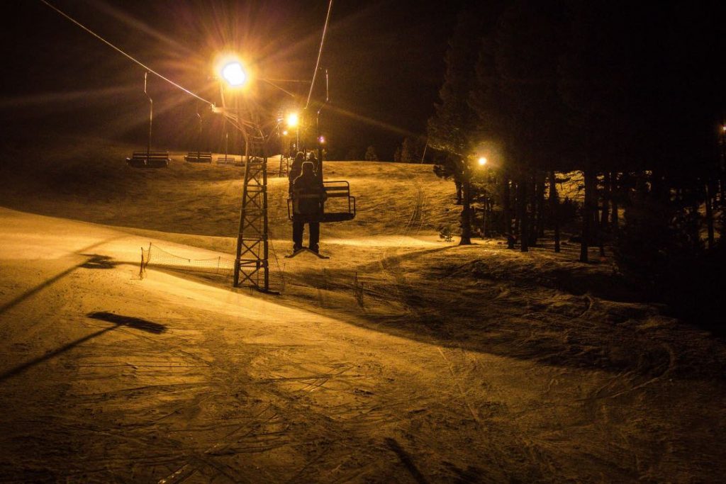 Double chairlift illuminated at night with a few skiers riding.