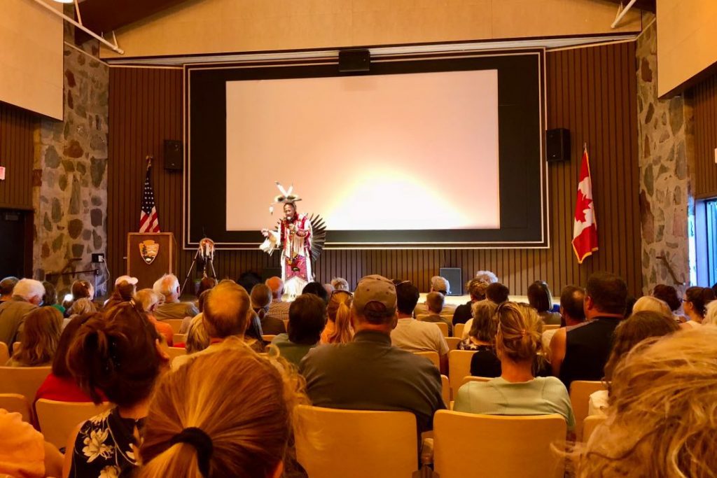 Native American speaker in traditional dress speaks to a packed auditorium.