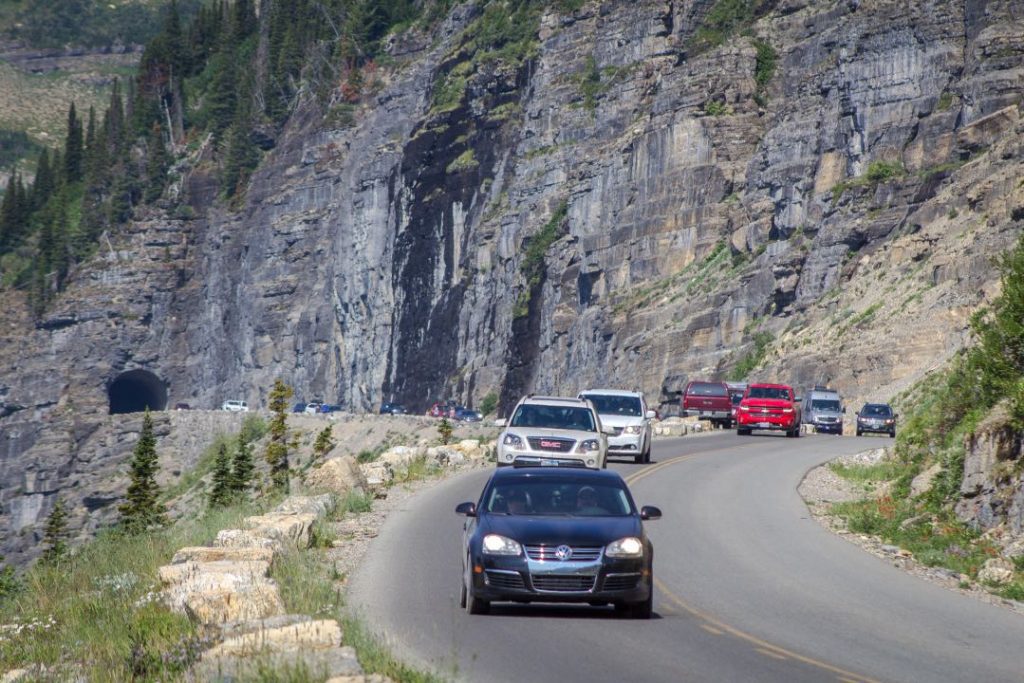 A line of cars winds its way through a narrow mountain road with a sheer rock wall on one side.
