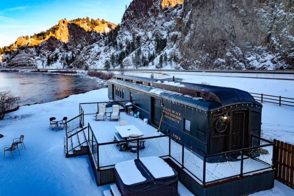 Parked train car with "Railway Express Agency" written on the side and a snow-covered patio sit in front of a rocky mountain and a body of water.