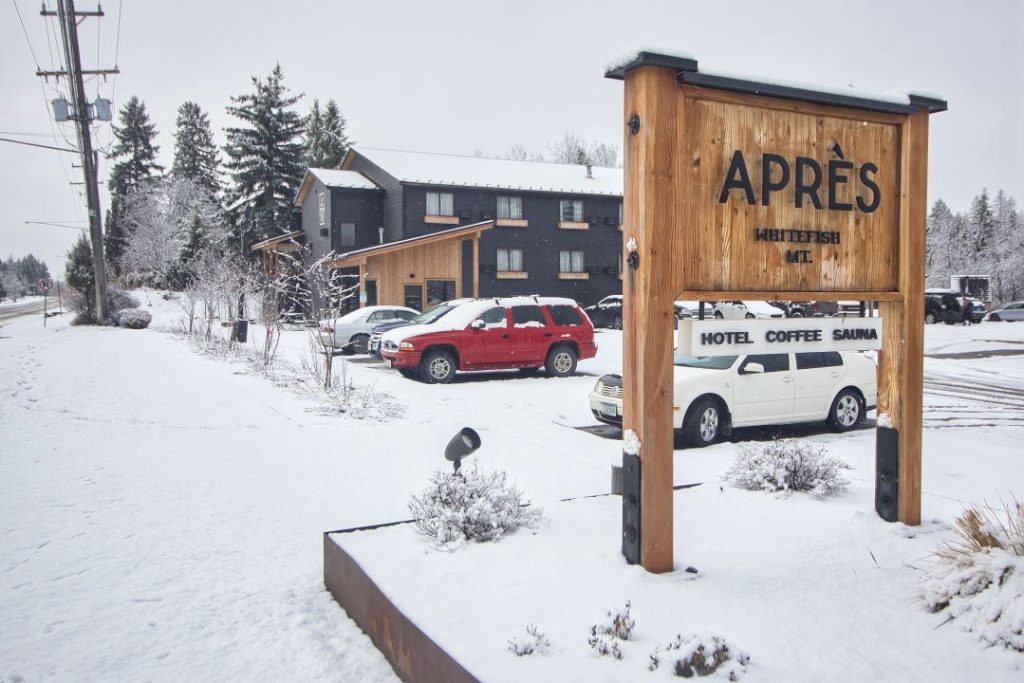 A wooden sign with black lettering says "Apres, Whitefish MT". Behind it is a two-story building with black siding and natural wood accents. The parking lot and surrounding lawn is covered in snow and several cars are in its parking lot.