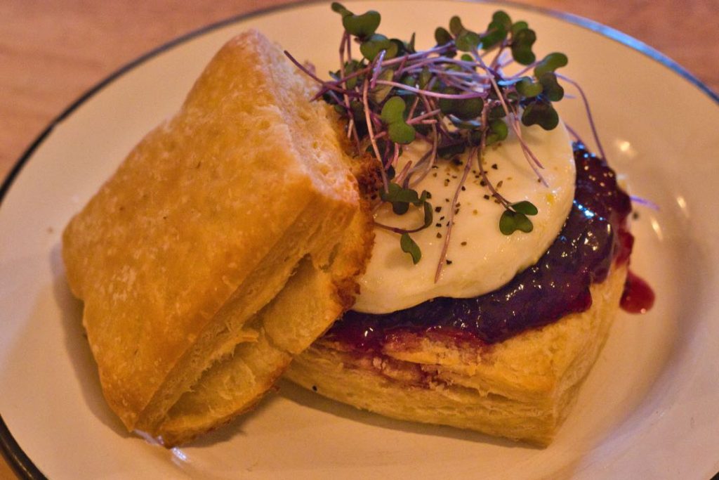Two fluffy biscuits on a plate. One is topped with a red jam, poached egg, and microgreens.