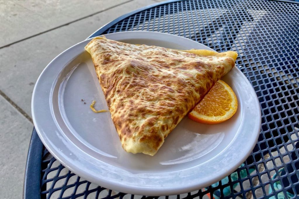 A crepe folded into a triangle on a white plate accompanied by an orange slice. The plate is sitting on a wrought iron table.