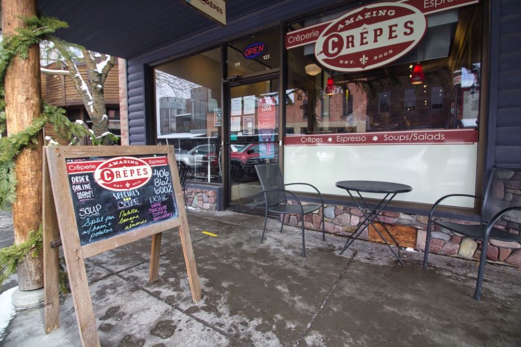 A chalkboard sandwich board sign with a multi-color menu on it sits outside a shop with a sign that says "Amazing Crepes" in the window. A wrought iron patio table and two chairs sits outside it.