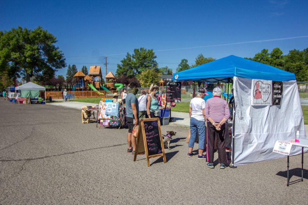 A chalkboard sign says "Pies" and lists off the flavors in front of a vendor tent. Several customers and a dog are ordering from it.