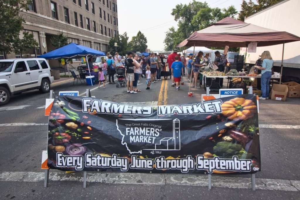 A black banner that says "Farmers Market - Every Saturday June Through September" with a logo of the outline of the state of Montana and a tall builinding inside of it. Behind the banner are people shopping at vendor tents on a closed off street.