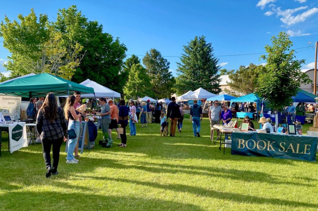 People milling about on a grassy field ringed with vendor tents. A table with a banner that says "Book Sale" is in the foreground.