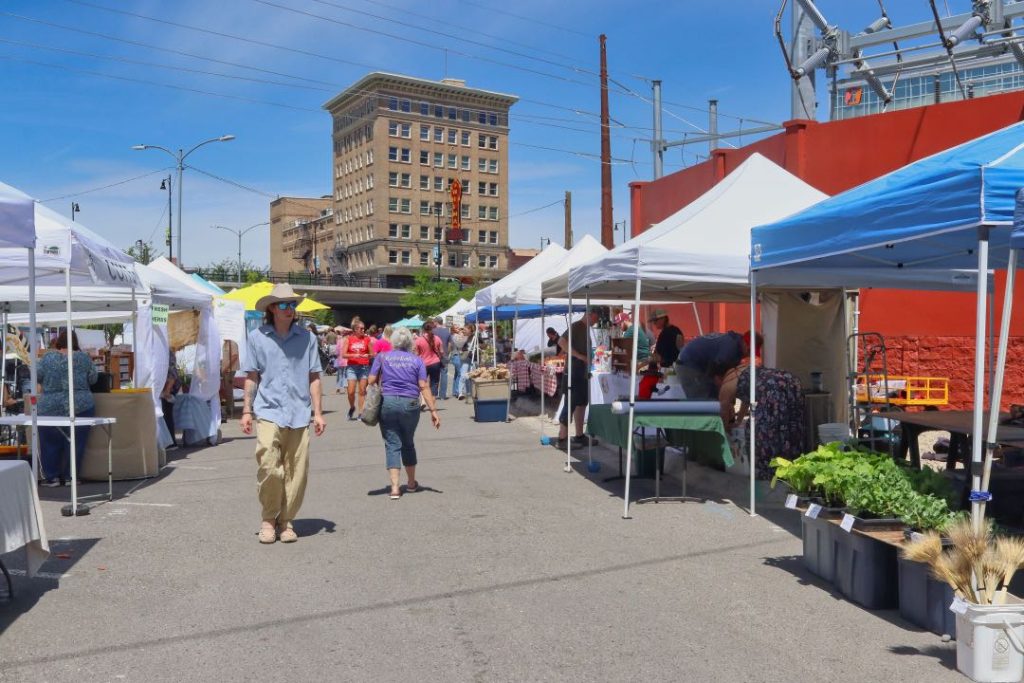 A man in a cowboy hat a blue button down shirt walks between rows of vendor tents at a farmers market.