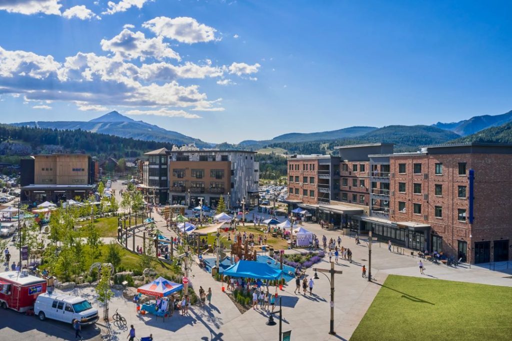 A mountain town with a green space populated with tents and farmers markets patrons. Four story brick buildings surround the green space.