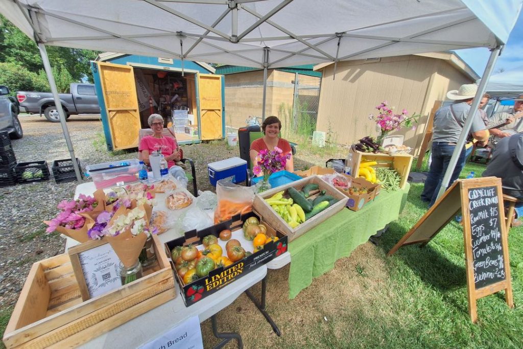 A women are seated behind a table with a green table cloth covered in produce and flowers for sale. Beside the table is a chalkboard sign with prices for the produce.