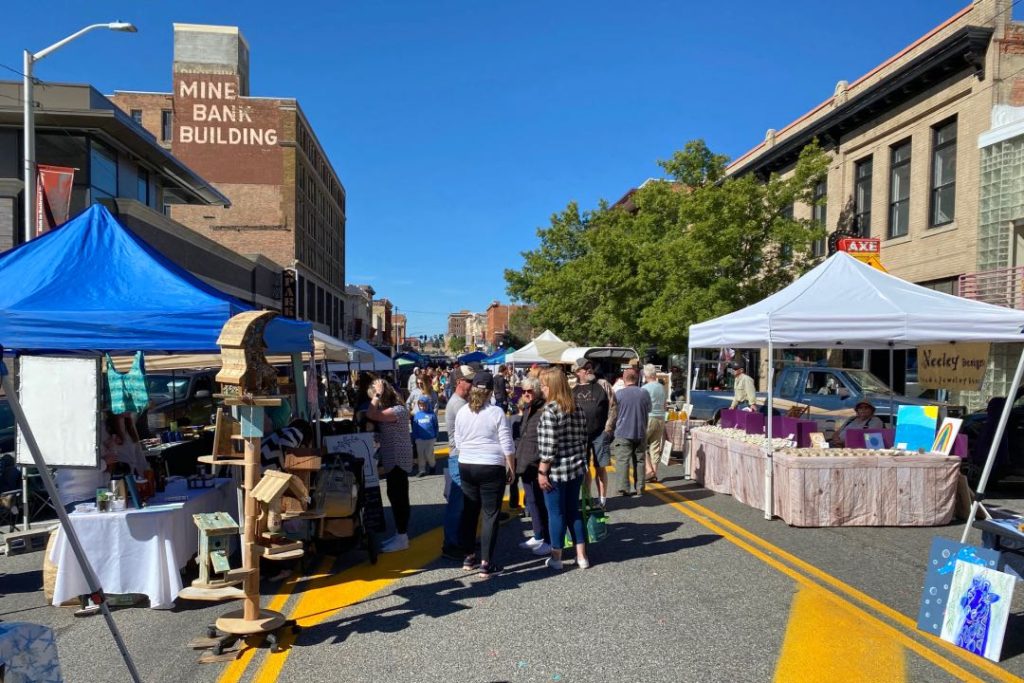 A city street lines with tents and tables for a farmers market. In the foreground is a display of wooden birdhouses.