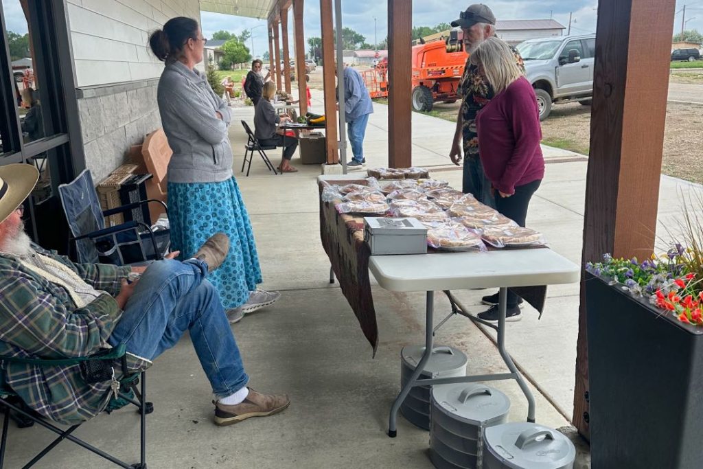 A bearded man in jeans and a wide brim hat sits in a camp chair behind a table of pies. A woman in a long blue skirt is selling these pies to a couple on the other side of the table.