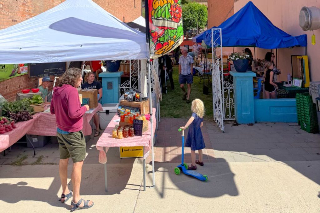 A man in shorts and sandals stands beneath an awning and next to a table stacked with jars of honey. A girl in a blue dress with a scooter looks past him at another set of tables and awnings.