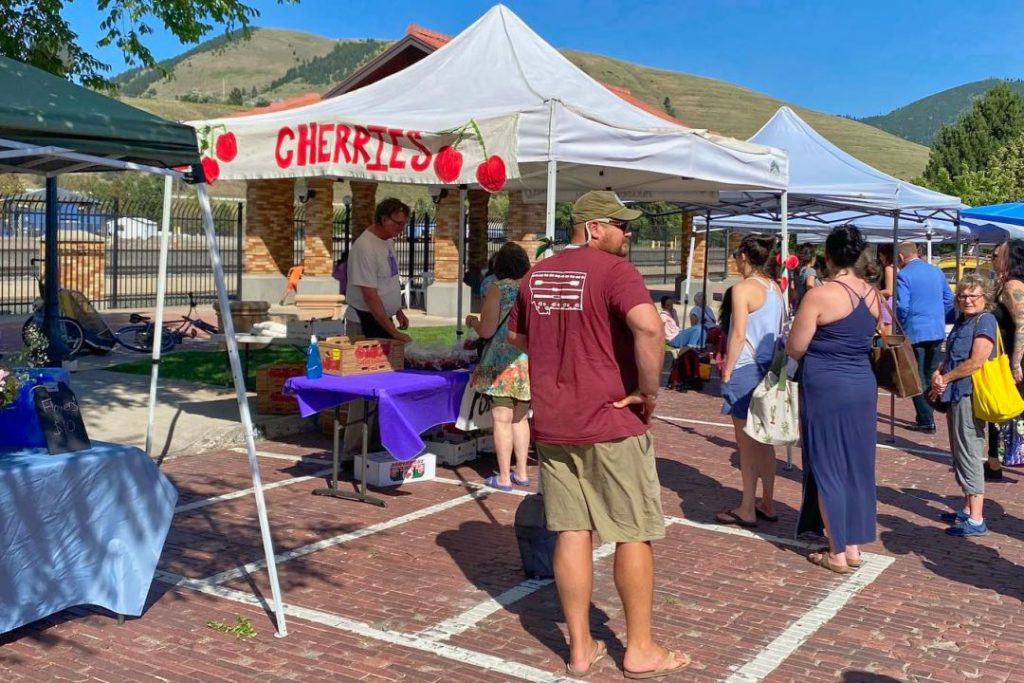 A man in a red t-shirt and shorts stands with several other customers in front of a table and awning selling cherries. A mountain is visible in the background.