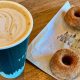 A paper coffee cup with a heart drawn in milk foams sits next to two small donuts covered in sugar.