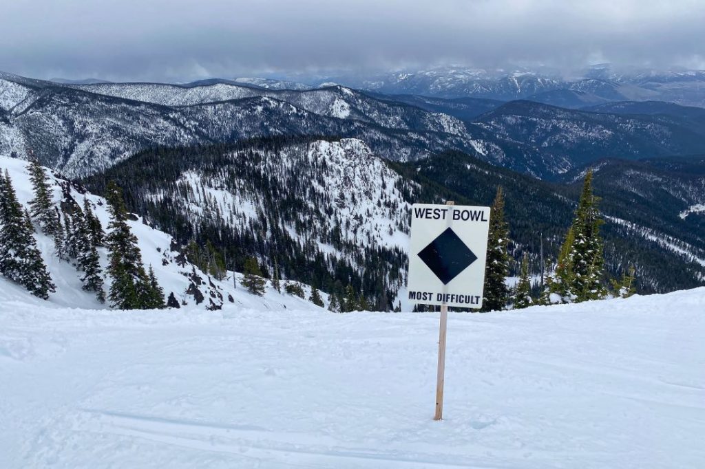 A white sign says "West Bowl" with a black diamond below it. It sits above a steep snowy hill with rugged-looking tracks from previous skiers and snowboarders.
