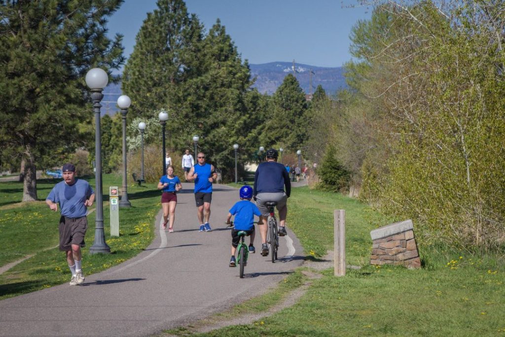 A father and sign ride bicycles on an asphalt path. Near them are several people jogging through the park area.