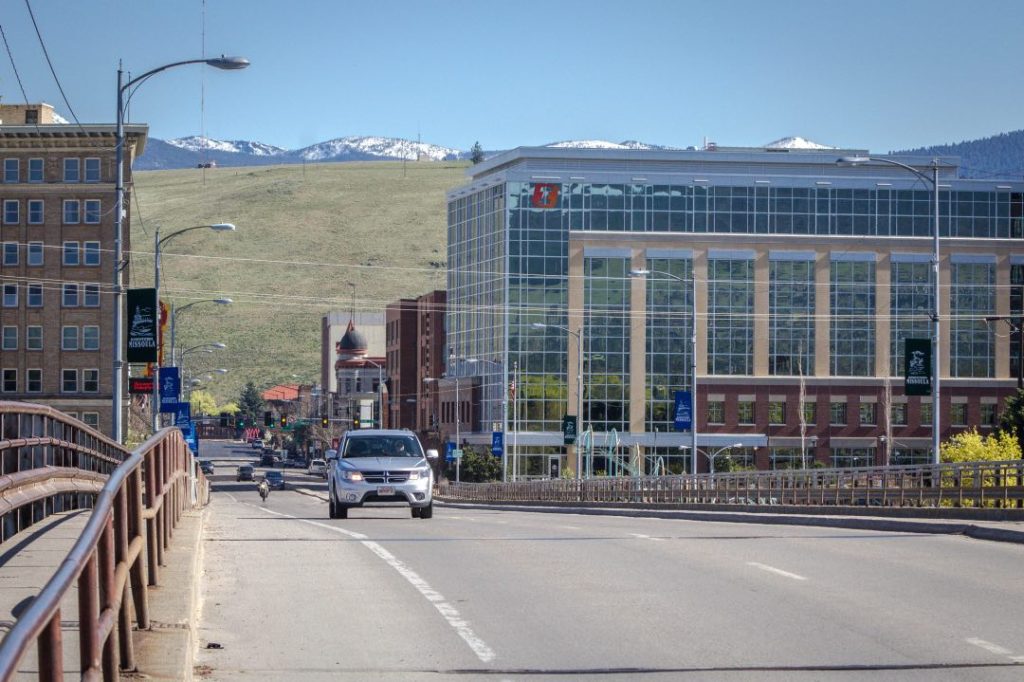 A car approaches over a bridge. In the background are several taller buildings.
