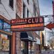 A red neon sign saying "Missoula Club, Burgers & Beer" with a red arrow pointing towards a storefront.