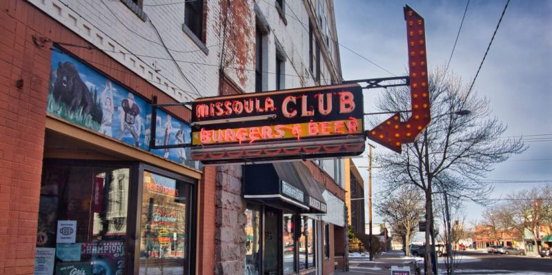 A red neon sign saying "Missoula Club, Burgers & Beer" with a red arrow pointing towards a storefront.