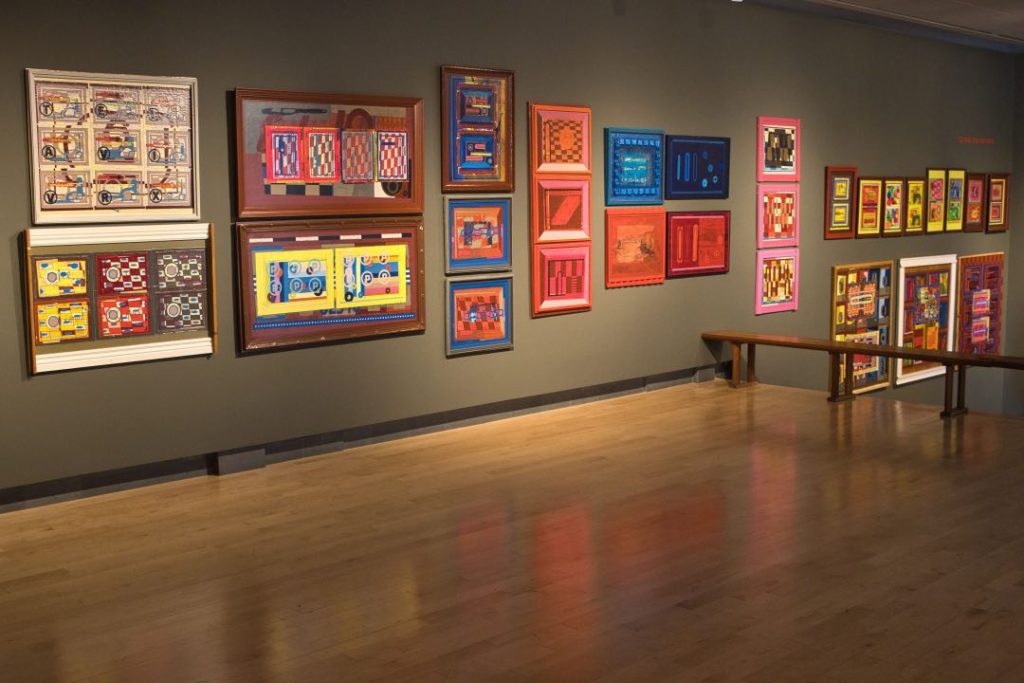 Dozens of abstract, geometric artworks are hung on a olive-colored gallery wall. A wooden floor is below them.