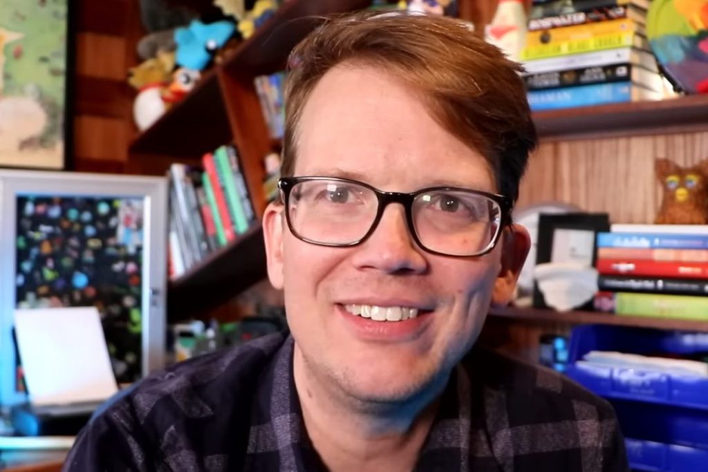 Photo of Youtuber Hank Green wearing glasses, smiling, and with an assortment of books in his background.