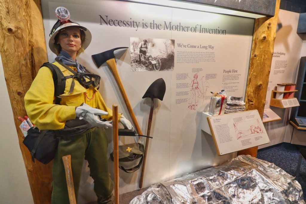 A female mannequin in yellow and green firefighting gear stands next to a set of tools including a shovel and axe.