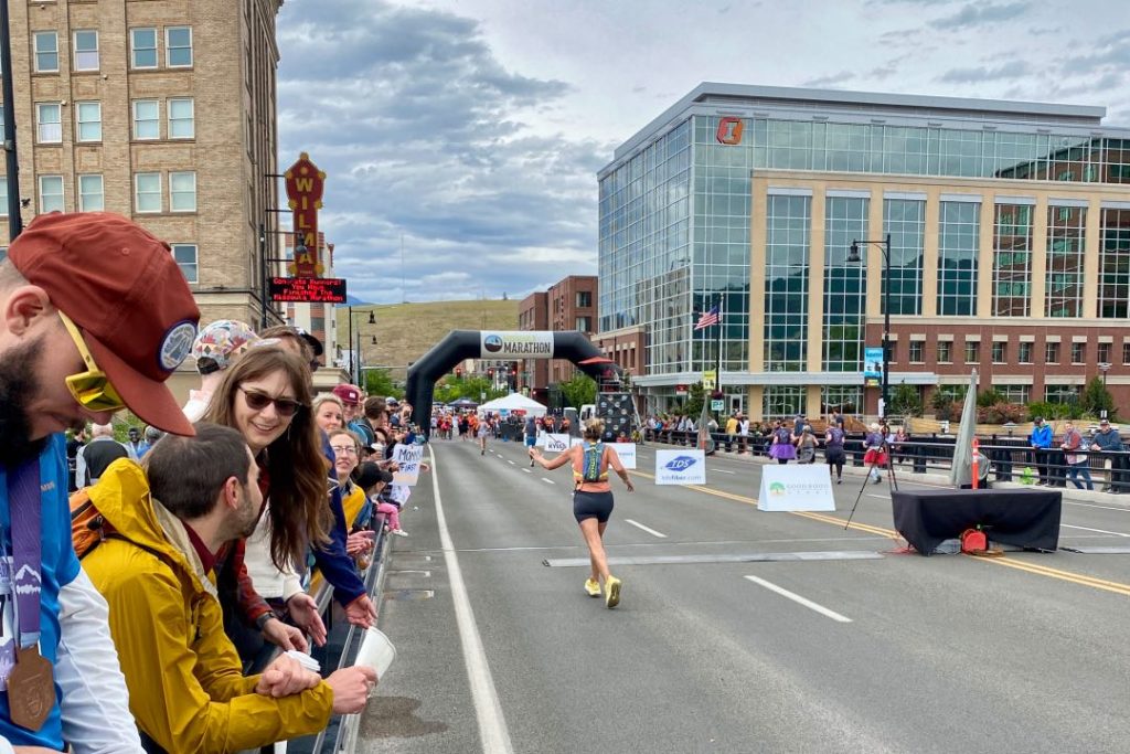 A woman seen from behind carrying a small backpack and running towards an inflatable finish line. Alongside her are hundreds of people cheering her on.