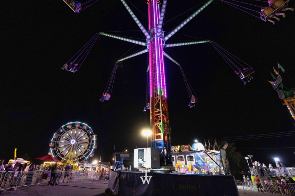 A carnival ride, lit up in purple, swings fairgoers in a circle, in front of a ferris wheel.