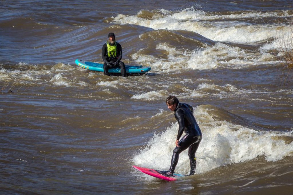 A man in a black wetsuit stands on a pink surfboard, riding a wave in the river. Behind him is a man in another black wetsuit, sitting on a blue surfboard.