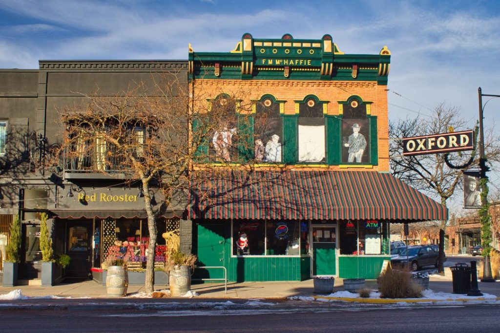 A green storefront with striped awning over it. A sign hangs from a light post nearby that says "Oxford". In the building's second floor windows are life-size drawings of historical characters.