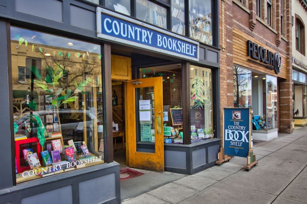 A bookstore with a blue sign above it's entryway that says "Country Bookshelf". On the sidewalk outside the store is a blue sandwich board-style sign that also says "The Country Bookshelf".