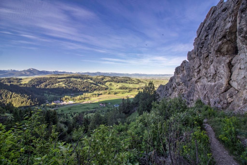 A narrow dirt path runs along a vertical rock wall. In the distance are rolling green hills and a nearly cloudless blue sky.
