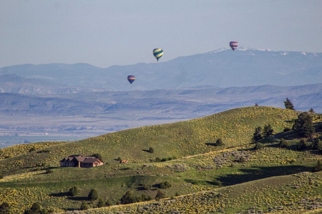 Three hot air balloons float above green foothills. Far off in the distance is a large mountain range.