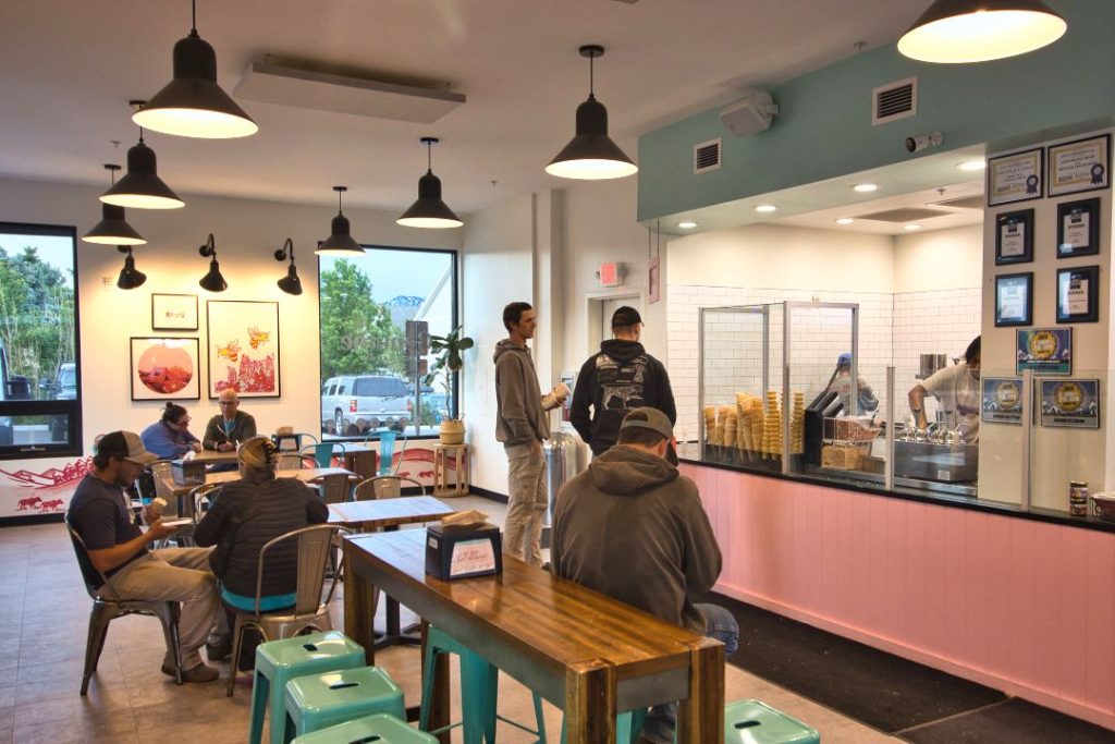 A group of customers sit on steel chairs and teal-painted metal stools. They're waiting for their order at pink ice cream counter which has a glass case of waffle cones on it.