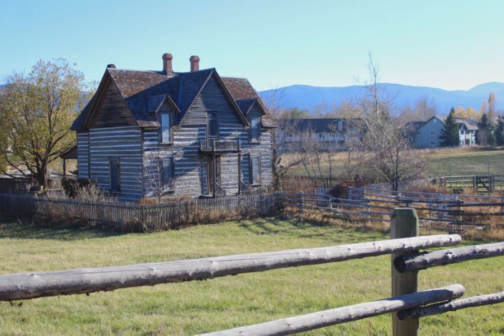 A pioneer era cabin sits behind an old picnic fence in a green pasture. In the distance are some modern homes and apartments.
