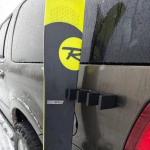 A black plastic ski holder is attached to the side of a grey SUV, holding a single black and yellow ski.