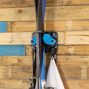 A black and blue wall mount for skis is attached to a wall made of unfinished wood. A pair of piles and a helmet bag also hang from the mount.