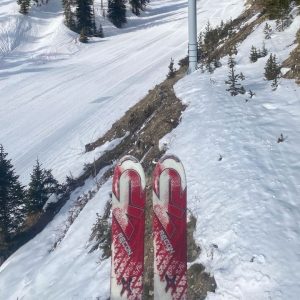 A pair of red and white skis as seen looking down from a chairlift. A snowy, but patchy slope is below.