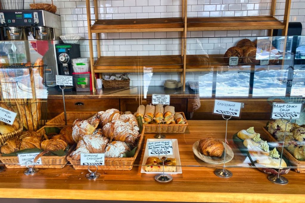 Baskets of croissants,scones, and wraps sit on a wooden counter behind a plexiglass sneeze guard. Behind it are mostly empty wooden shelves, with a few loaves of artisan bread.