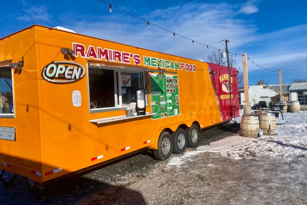 An orange and red food truck labeled "Ramire's Mexican Food" sits in a snowy parking lot with a string of lights draped over the order window.