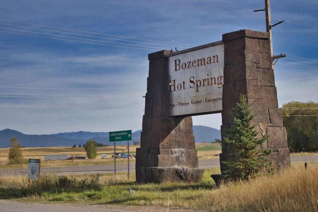 A large stone sign says "Bozeman Hot Springs". Behind it is an open prairie, and in the distance, a small mountain range.