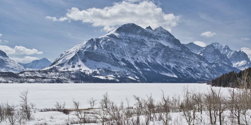 A frozen and snow-covered lake sits beneath an angular mountain, also covered in snow.