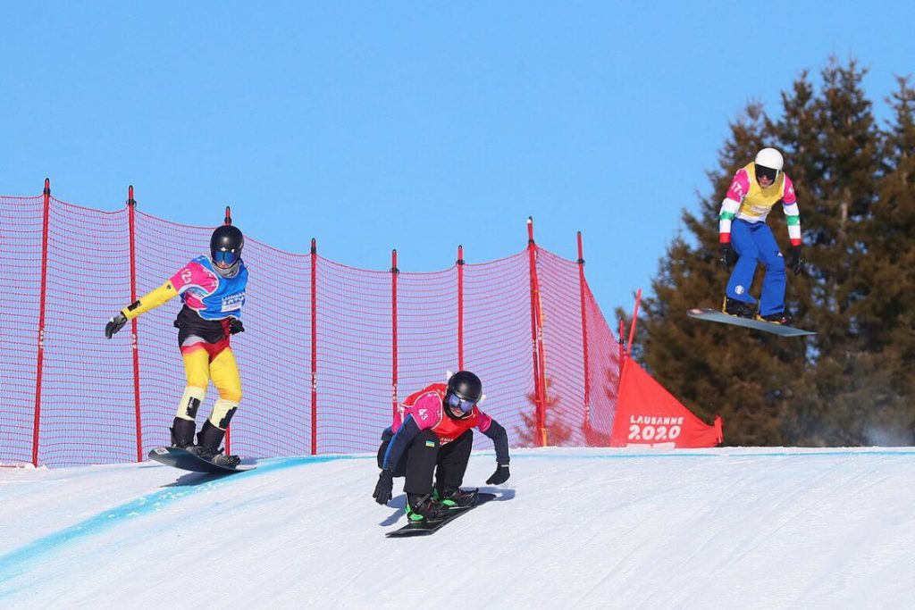 Three snowboarders in competition racing bibs compete to get down a race course.