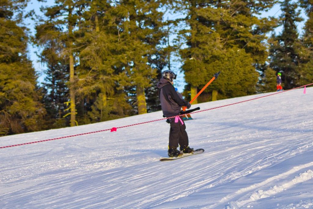 A snowboarder in black pants, dark jacket, and helmet, rides a t-bar lift up a groomed ski hill.