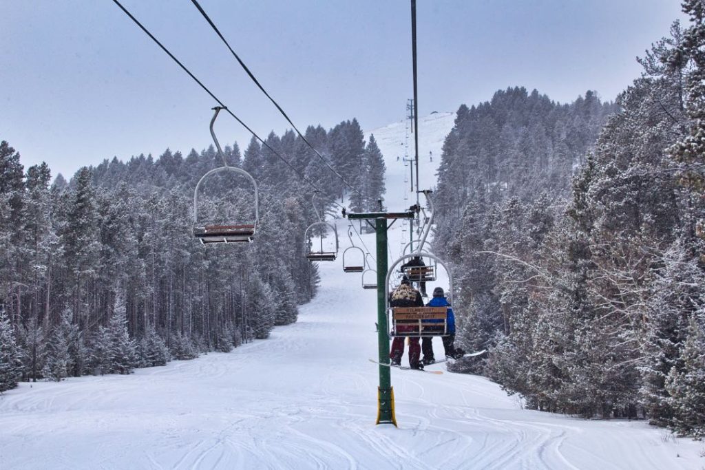 A pair of snowboarders ride a double chairlift up a run flanked by snow-covered trees.