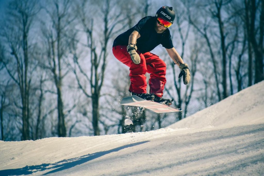 A man in red pants and a t-shirt jumps a snowboard.