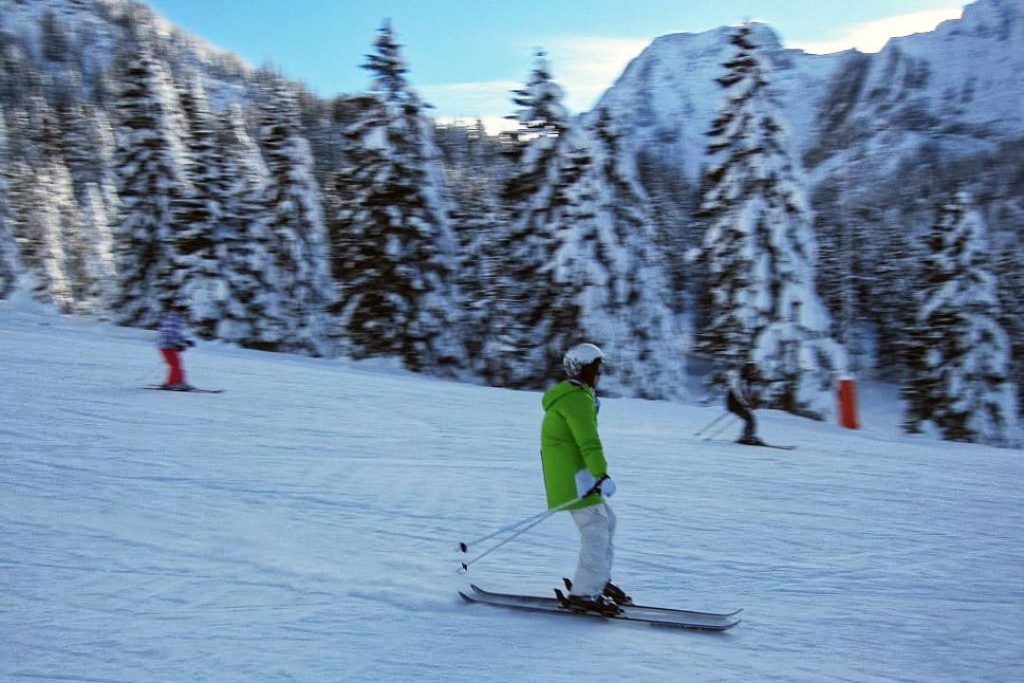 A skier in a green coat and white boards rides down a slope on twin tip skis. The background is heavily blurred.